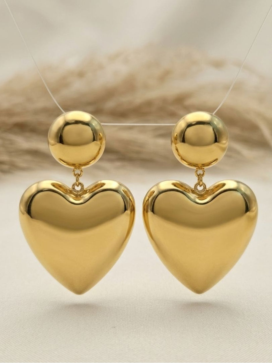 Gold heart drop earrings suspended on a display stand against a neutral linen background