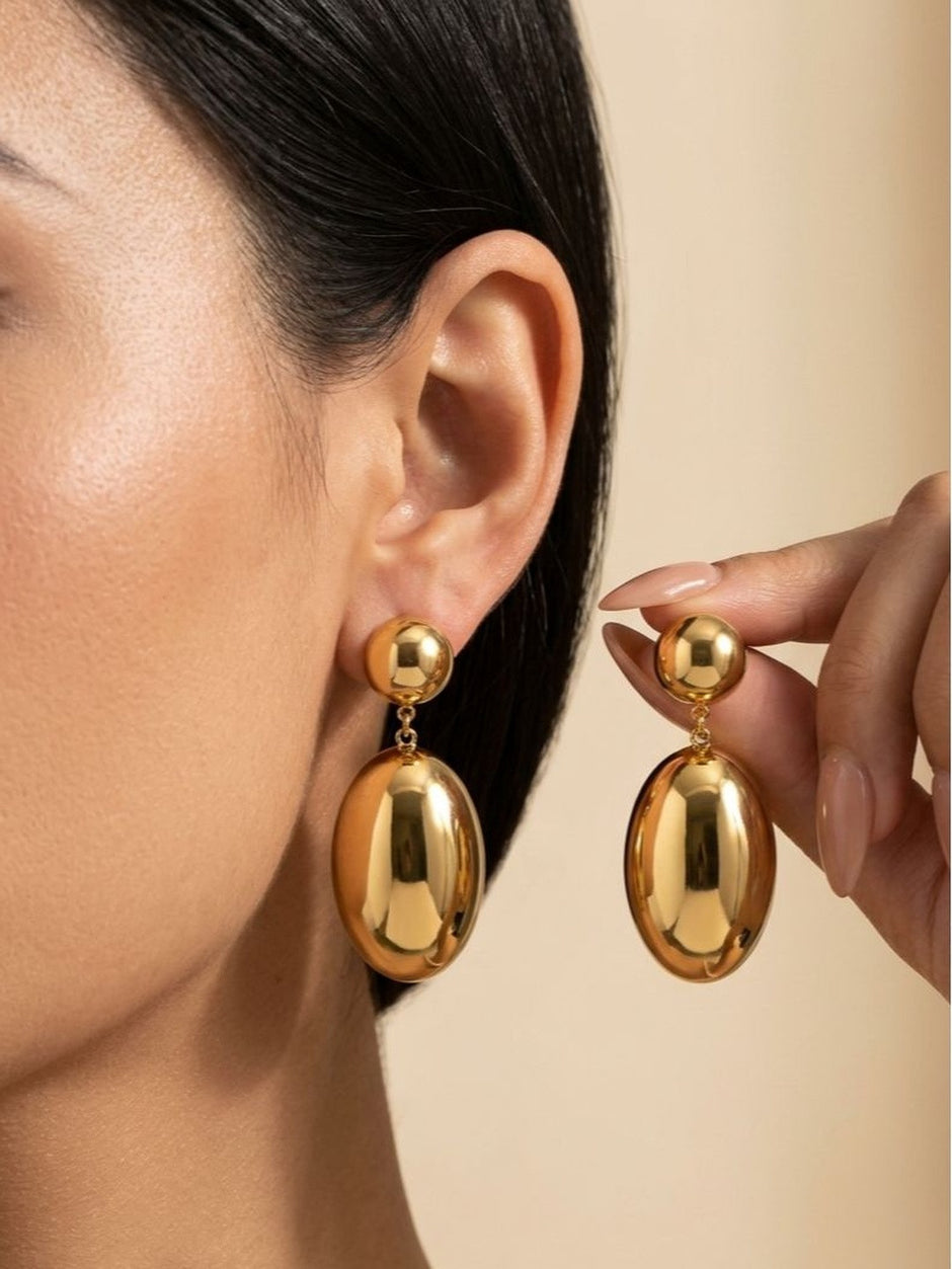 Woman wearing one gold oval drop earring while holding the second earring up beside her ear