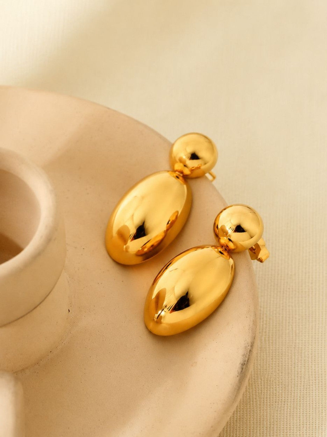 Two gold-plated oval drop earrings laid flat on a cream ceramic dish from above