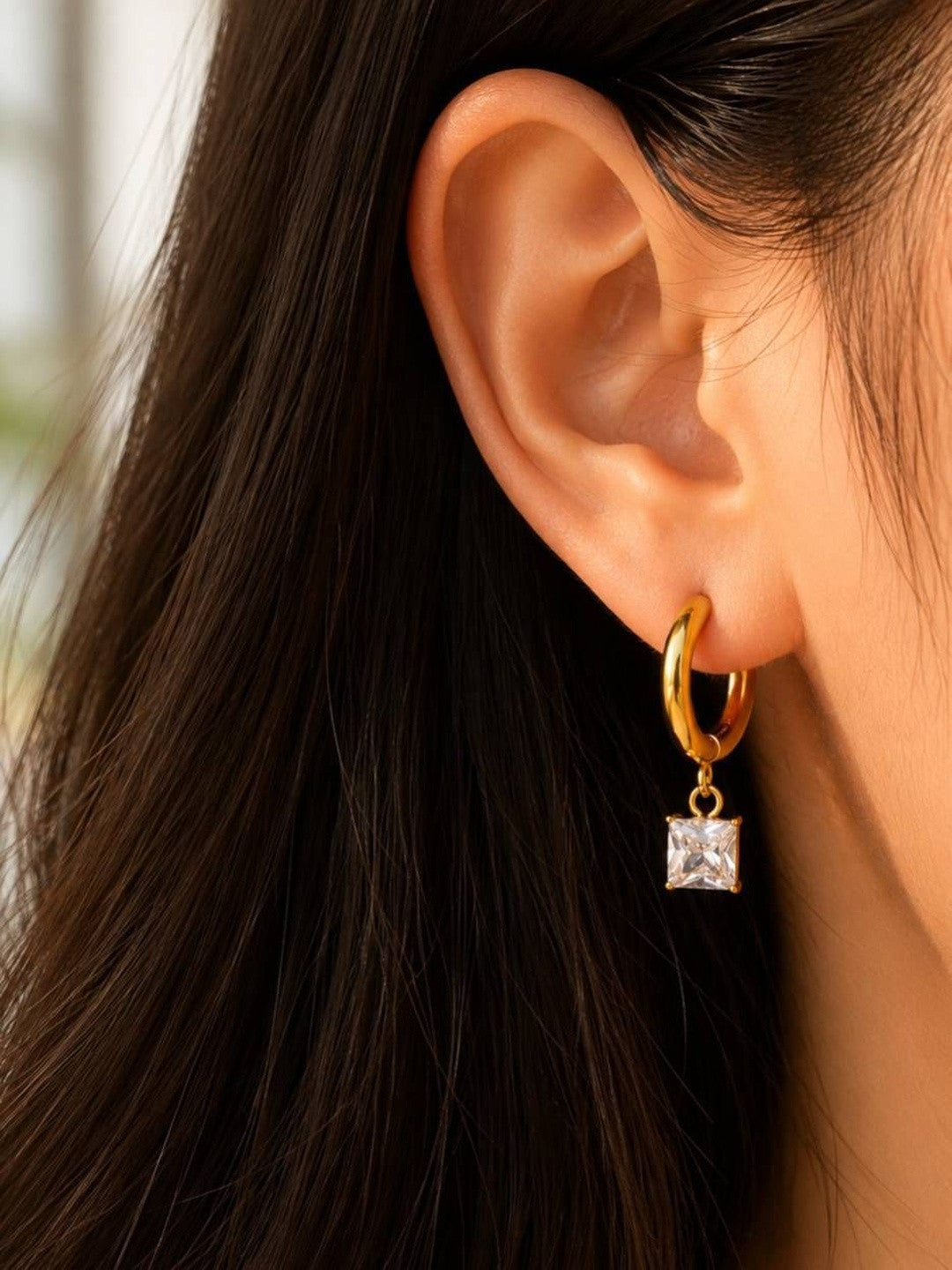 Model wearing a single gold huggie hoop earring with dangling square zirconia charm, dark hair backdrop
