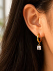 Model wearing a single gold huggie hoop earring with dangling square zirconia charm, dark hair backdrop