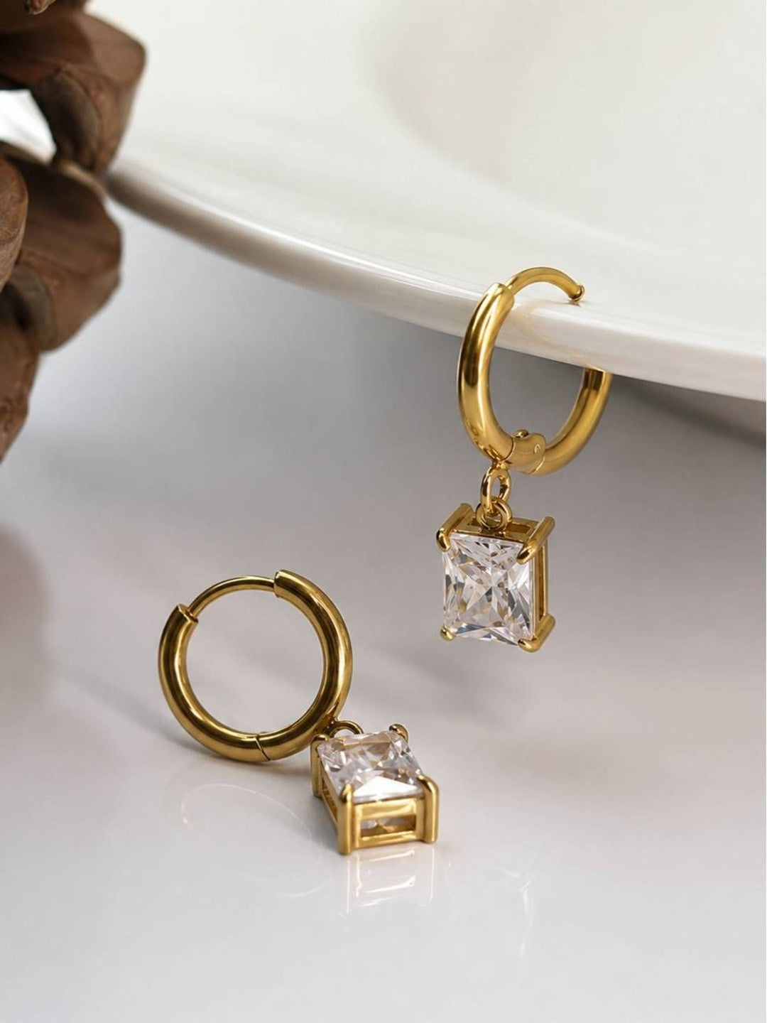 Two gold hoop earrings with square crystal drop charms resting on a white ceramic dish with pinecone beside