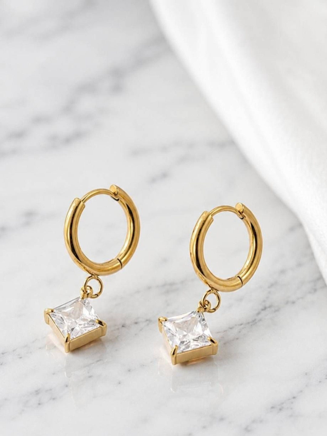 Gold huggie hoop earrings with square zirconia charms laid flat on white marble surface