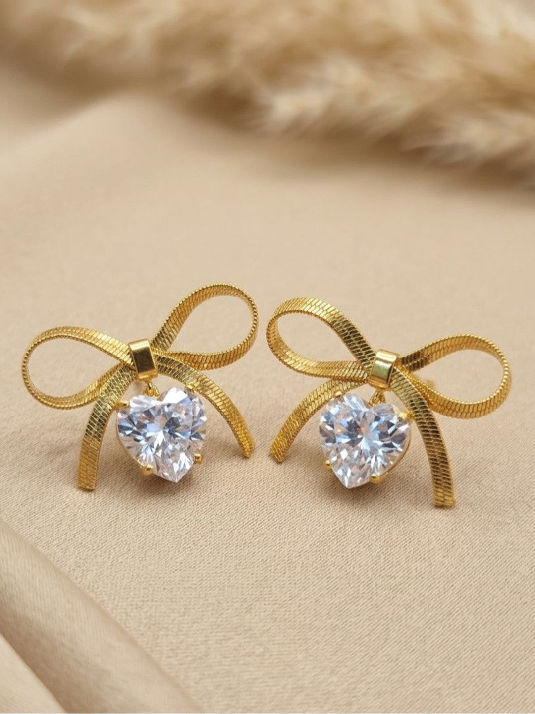 Pair of gold-plated herringbone bow stud earrings with heart-shaped cubic zirconia drops on a beige surface