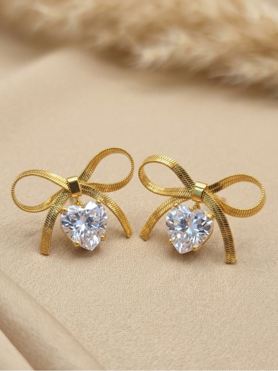 Pair of gold-plated herringbone bow stud earrings with heart-shaped cubic zirconia drops on a beige surface