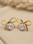 Pair of gold-plated herringbone bow stud earrings with heart-shaped cubic zirconia drops on a beige surface