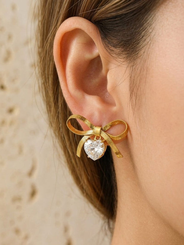 Gold bow stud earring with heart crystal drop worn on a woman's ear showing scale and fit