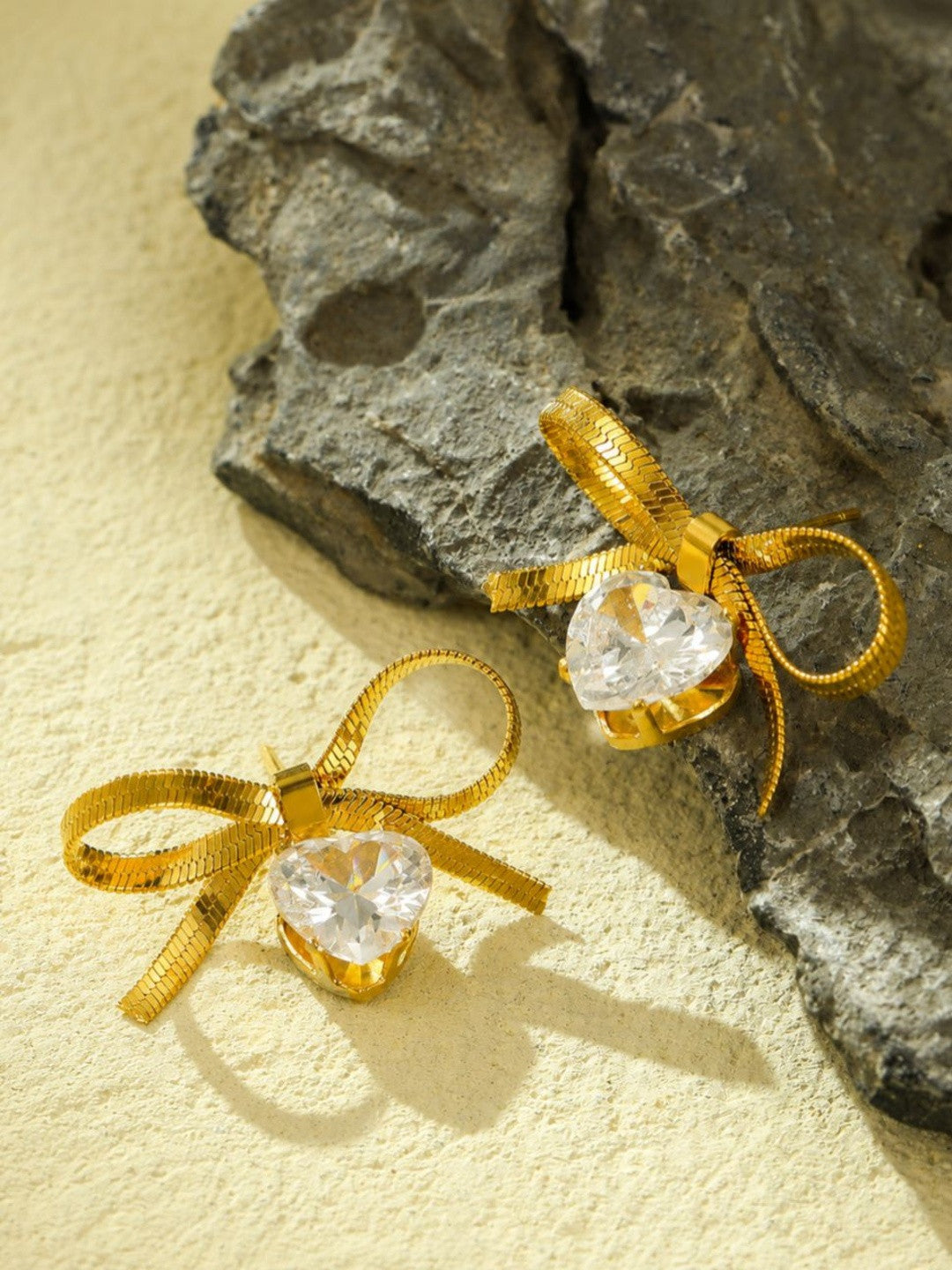 Two gold herringbone bow earrings with sparkling heart drops resting on a textured stone surface
