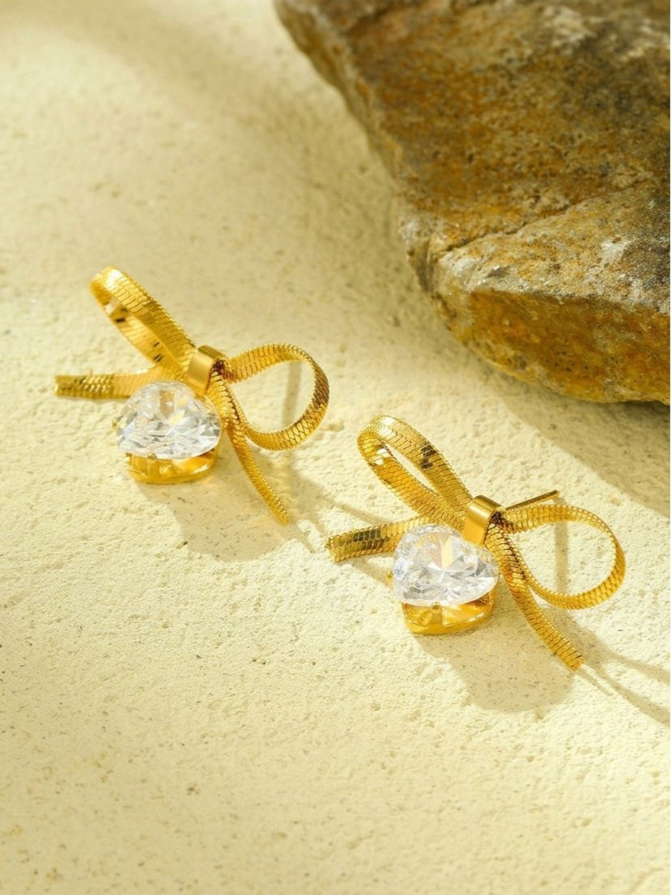 Close-up of gold bow stud earring pair with clear heart cubic zirconia drops on a sandy surface