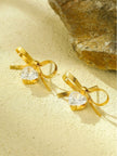 Close-up of gold bow stud earring pair with clear heart cubic zirconia drops on a sandy surface