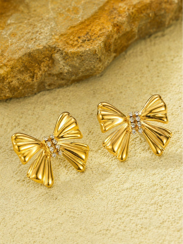 Pair of gold-plated ribbed bow stud earrings with crystal centre bar on sandy surface