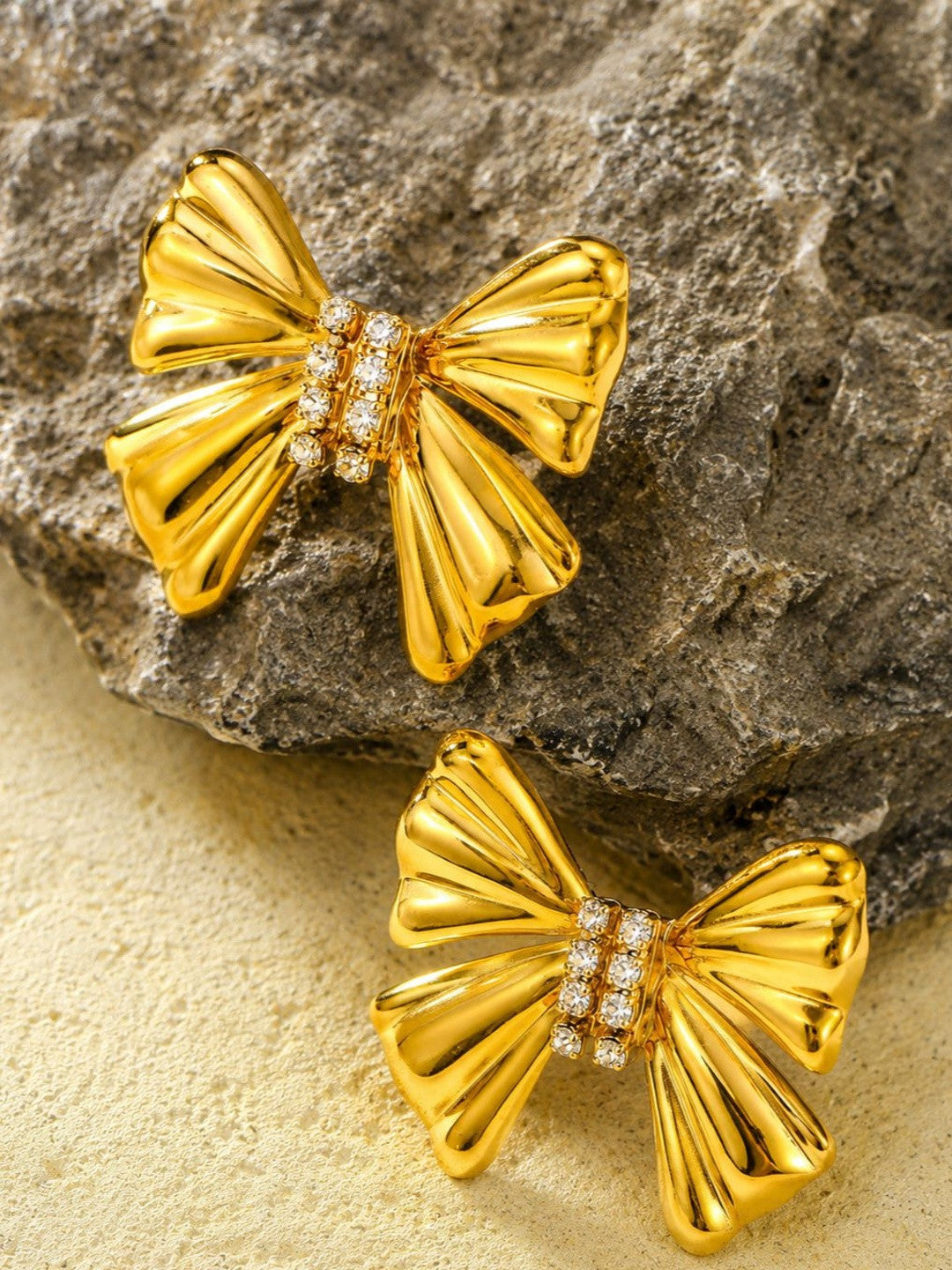 Two gold-plated bow stud earrings with crystal detail resting on a textured grey rock