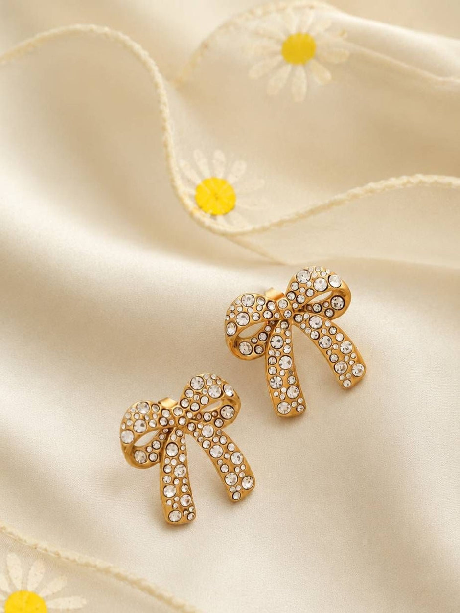 Pair of gold-toned crystal pavé bow stud earrings laid flat on cream daisy fabric