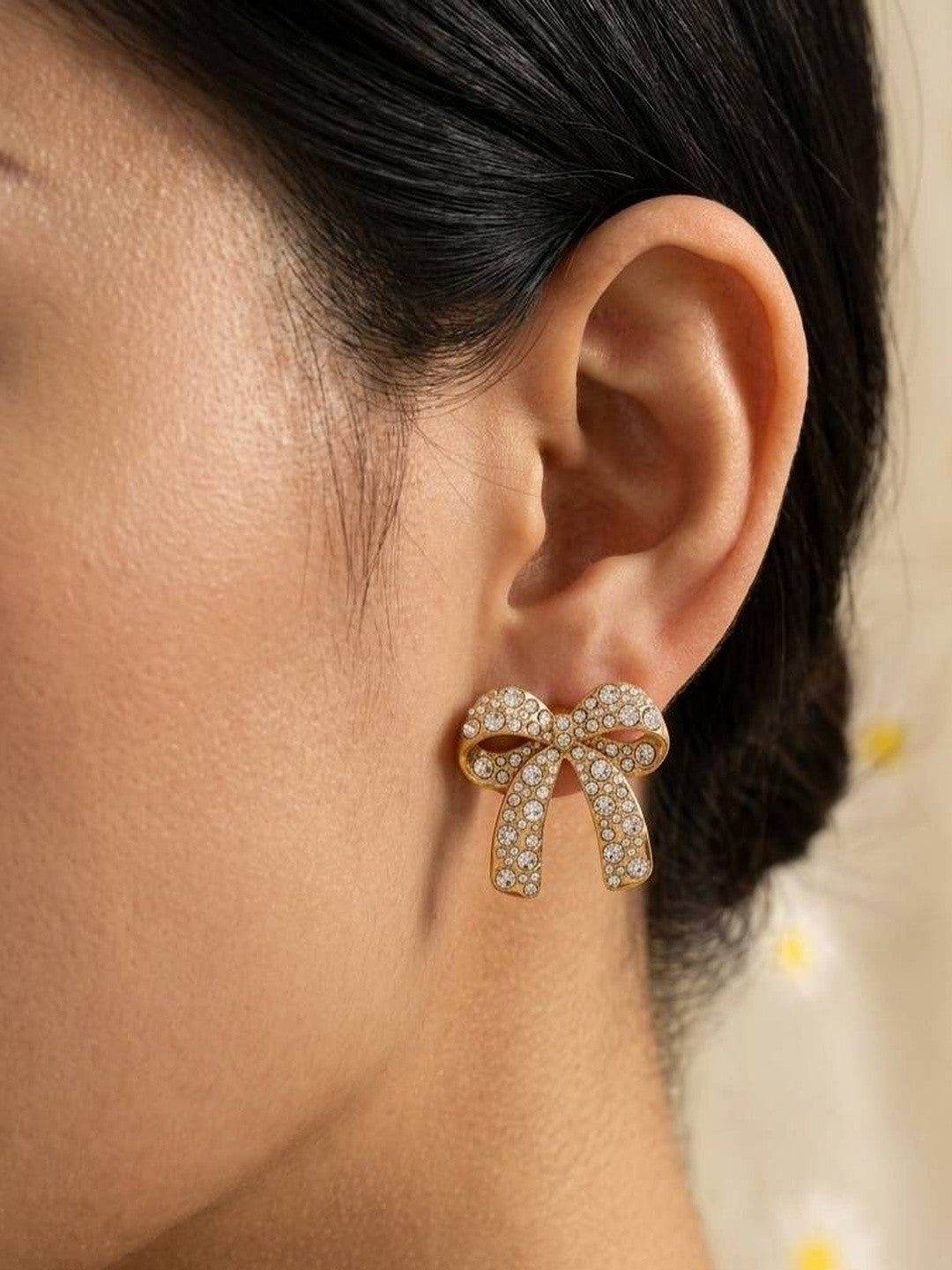 Side profile view of crystal pavé gold bow earring on model's ear with daisy background
