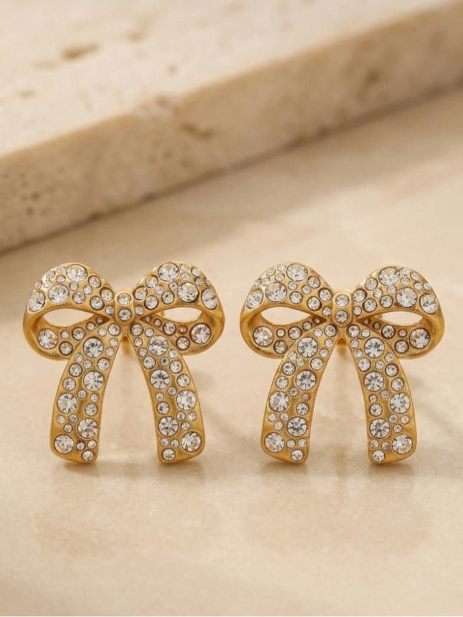Two gold-toned crystal bow stud earrings placed side by side on a stone surface