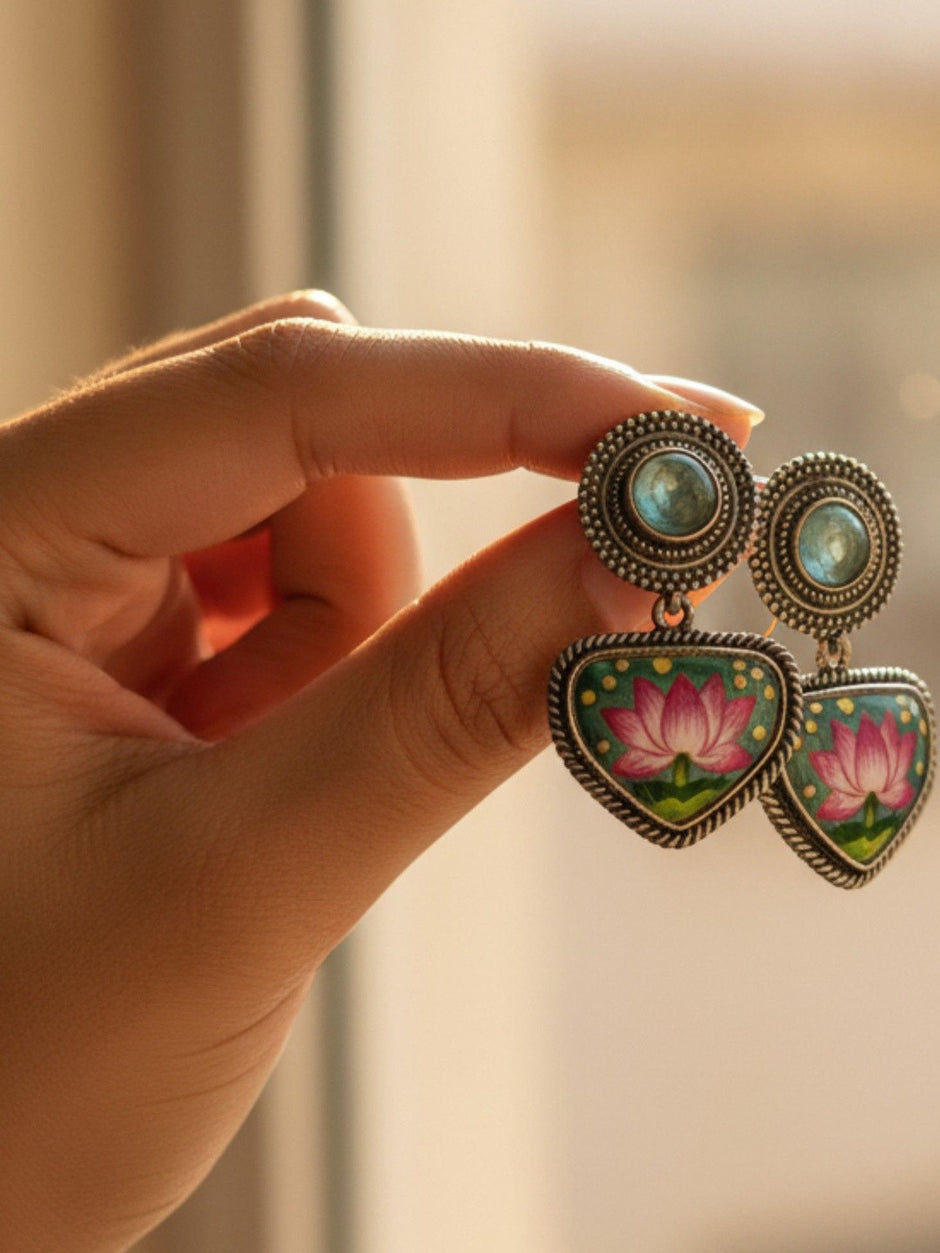 Hand holding pair of oxidised silver hand-painted lotus enamel drop earrings in warm natural light