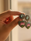 Hand holding pair of oxidised silver hand-painted lotus enamel drop earrings in warm natural light