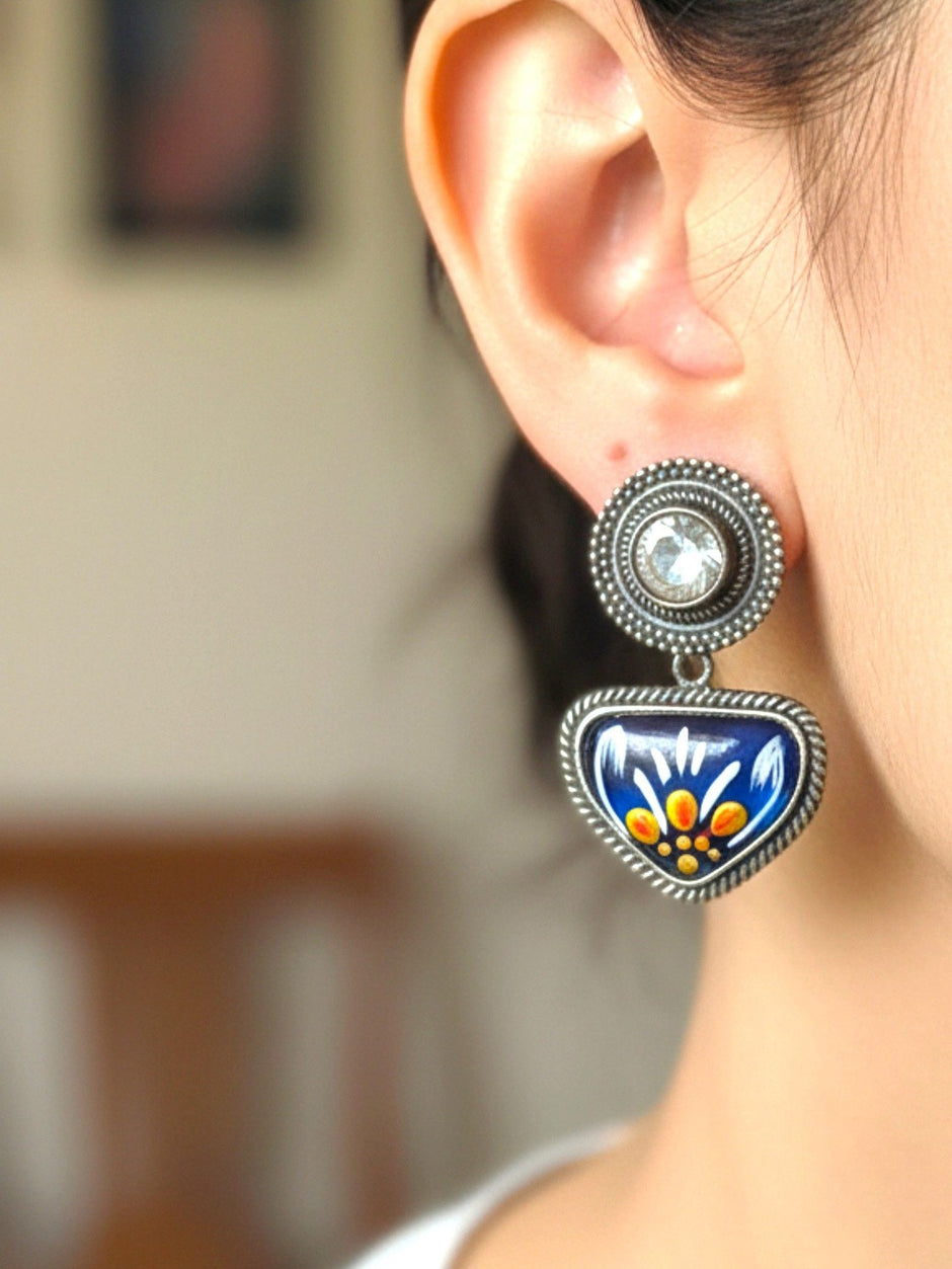Oxidised silver meenakari drop stud earring worn on a woman's ear, showing blue enamel pendant with floral motif and crystal stud top