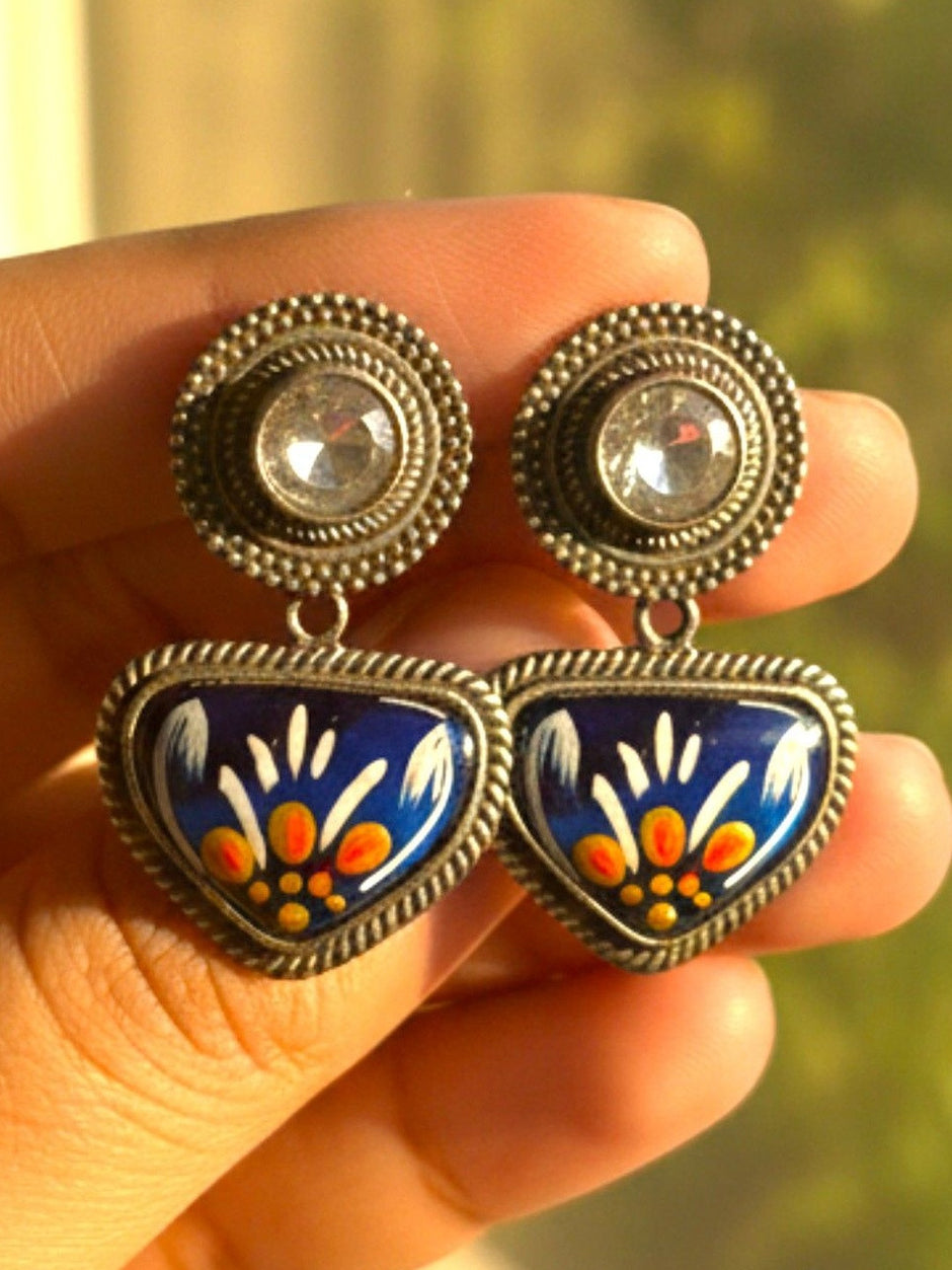 Pair of oxidised silver meenakari drop studs held in hand, displaying blue hand-painted enamel triangular pendants and crystal solitaire tops