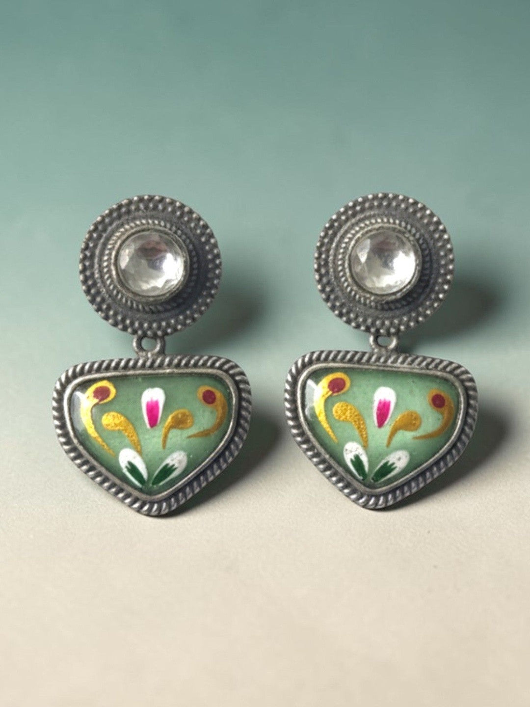Pair of oxidised silver meenakari studs with sage green enamel triangular drops on teal background