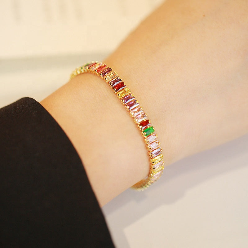 Model wearing a gold tennis bracelet with multicolored baguette-cut stones.