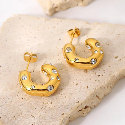 A pair of gold-tone hoop earrings with clear rhinestones or cubic zirconia embedded throughout the surface, displayed on a beige stone.