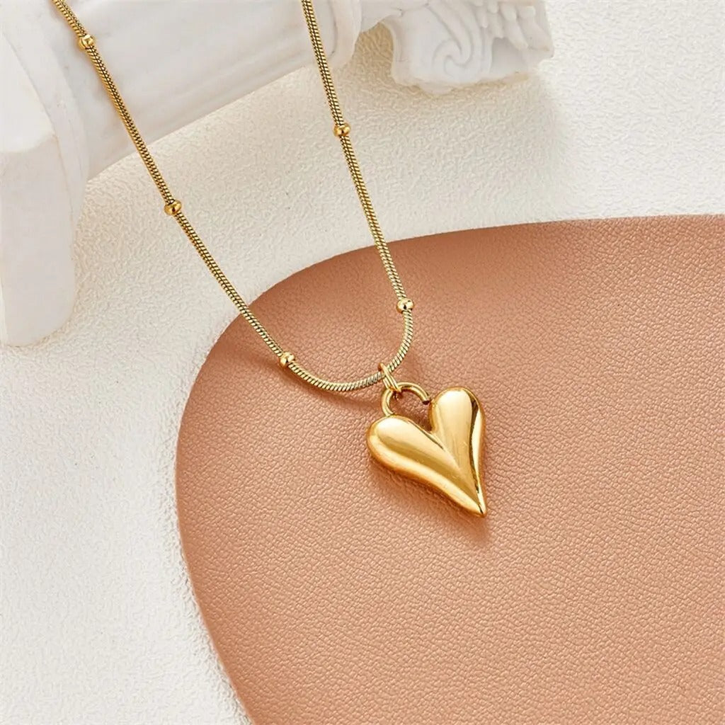 A gold heart pendant necklace with a beaded chain displayed on a white and gold surface.