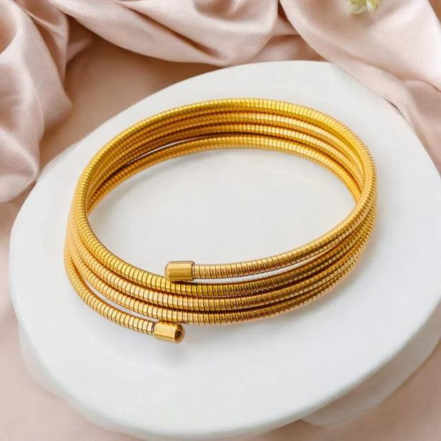 A gold-colored multi-layered spiral bracelet displayed on a white platform with a pink backdrop.