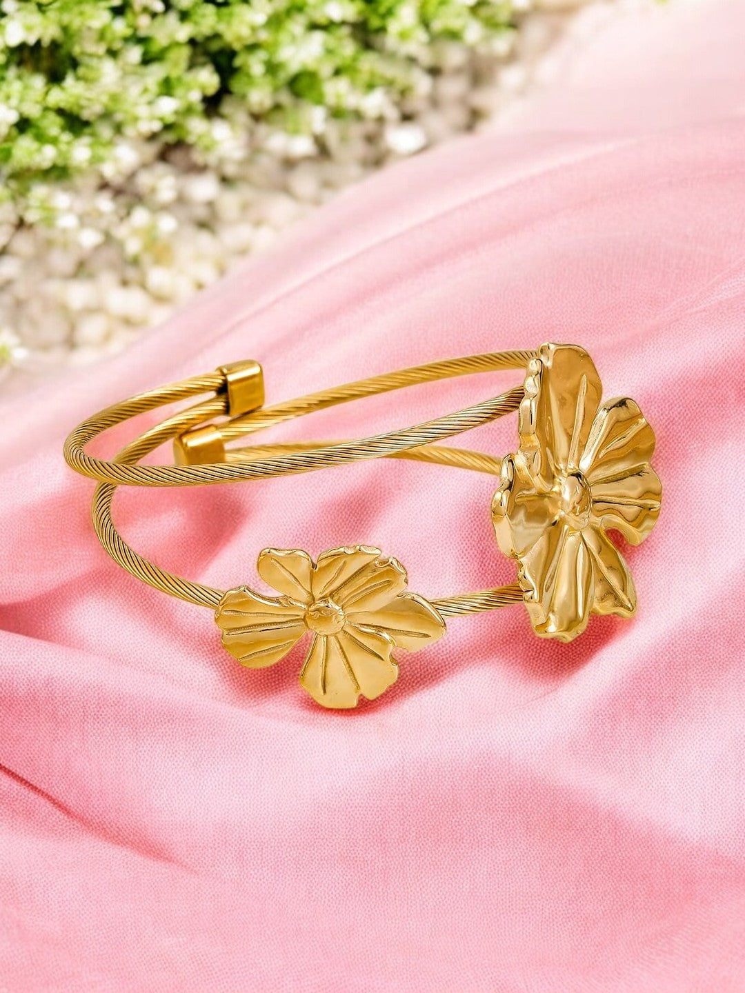 A gold-colored cuff bracelet with two flower designs displayed on a pink fabric surface.